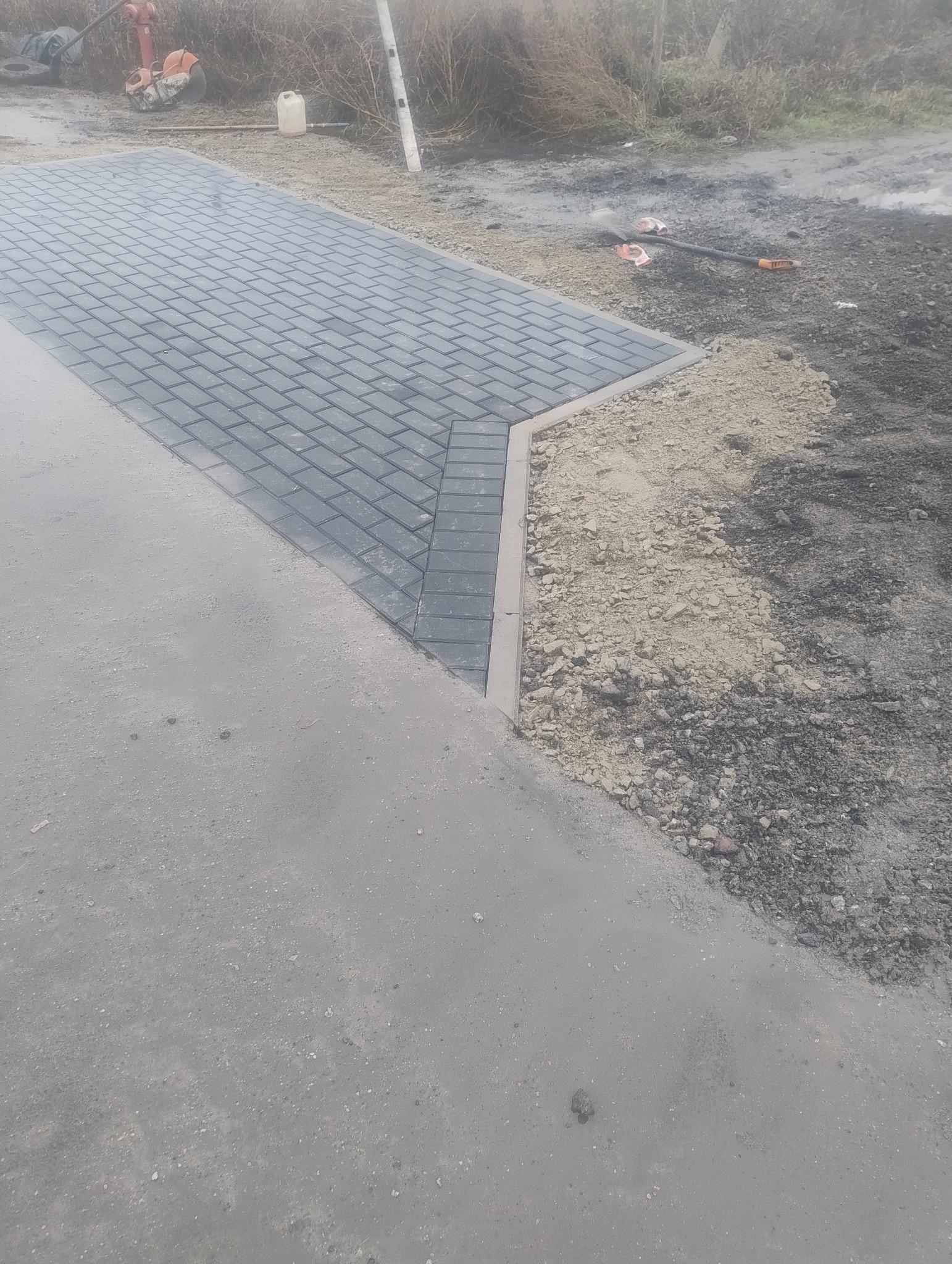 Układanie szarej kostki brukowej w jodełkę, widoczne narzędzia i przygotowane podłoże.