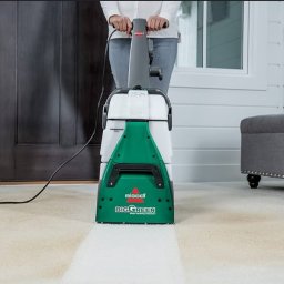 Silver Clean Maciej Borysławski - Kobieta czyści jasny dywan odkurzaczem piorącym Bissell Big Green, widoczny efekt czyszczenia w postaci jaśniejszego pasa na dywanie. W tle szara pufa i białe panele.