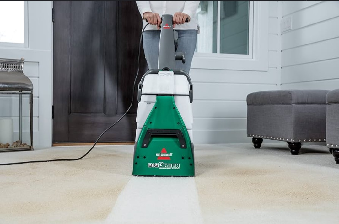 Kobieta czyści jasny dywan odkurzaczem piorącym Bissell Big Green, widoczny efekt czyszczenia w postaci jaśniejszego pasa na dywanie. W tle szara pufa i białe panele.