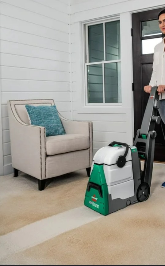 Kobieta czyści dywan maszyną Bissell, widoczny efekt czyszczenia w postaci jaśniejszego pasa na dywanie. W tle fotel i białe panele ścienne.