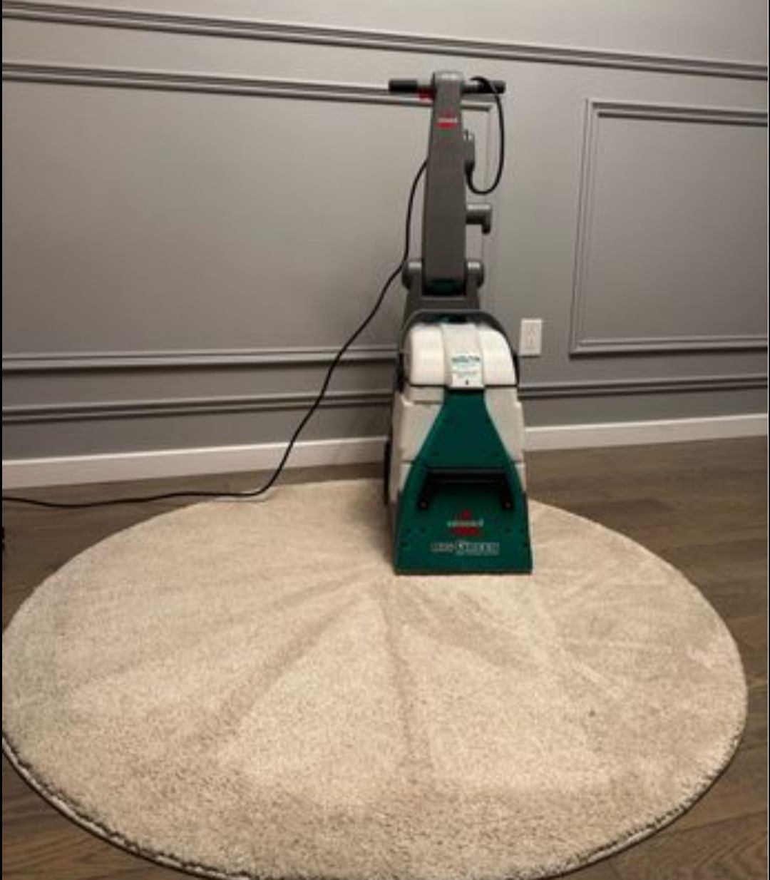 Okrągły, beżowy dywan po czyszczeniu, z widocznym urządzeniem do prania dywanów Bissell, na tle szarej ściany z dekoracyjnymi panelami.