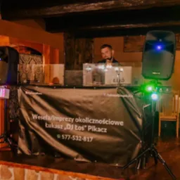 Stanowisko DJ-a Łukasza 'DJ Łoś' Pikacza z nagłośnieniem i oświetleniem na tle rustykalnego wnętrza z drewnianym stropem. Na banerze reklamowym widnieją dane kontaktowe i oferta: wesela/imprezy...