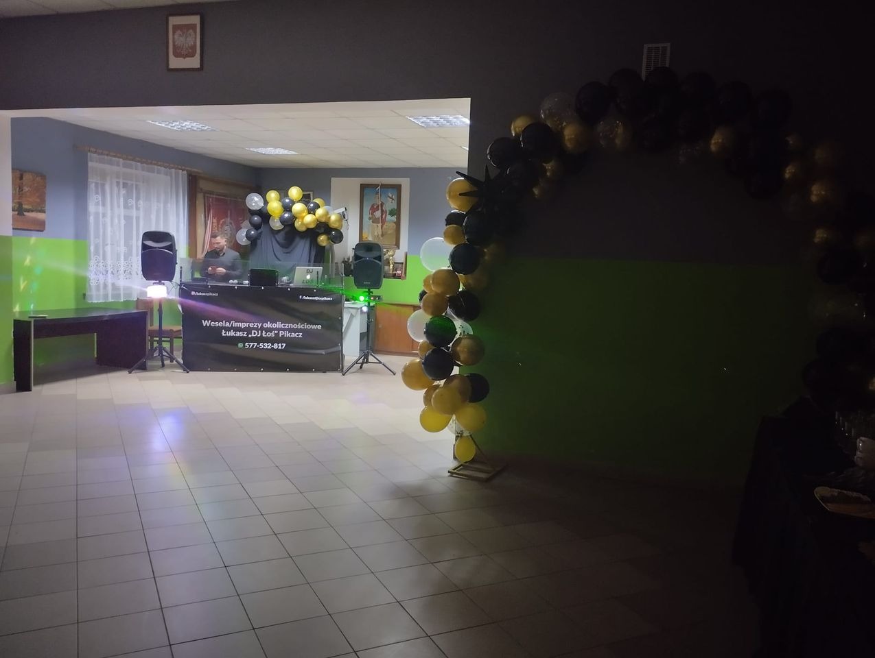 DJ Łukasz 'Pikacz' za konsoletą na tle dekoracji z czarno-złotych balonów, sala z parkietem w jasnych płytkach.