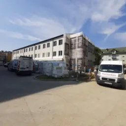 Prace na elewacji budynku wielorodzinnego w trakcie realizacji, zabezpieczone siatką ochronną z reklamą, palety styropianu obok, zaparkowane firmowe busy Remontex.