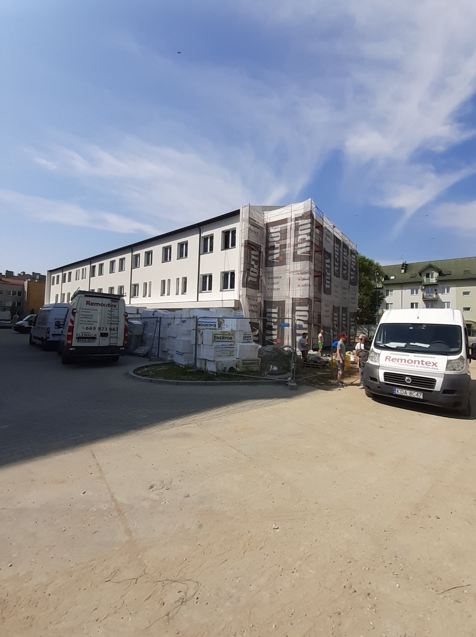Prace na elewacji budynku wielorodzinnego w trakcie realizacji, zabezpieczone siatką ochronną z reklamą, palety styropianu obok, zaparkowane firmowe busy Remontex.