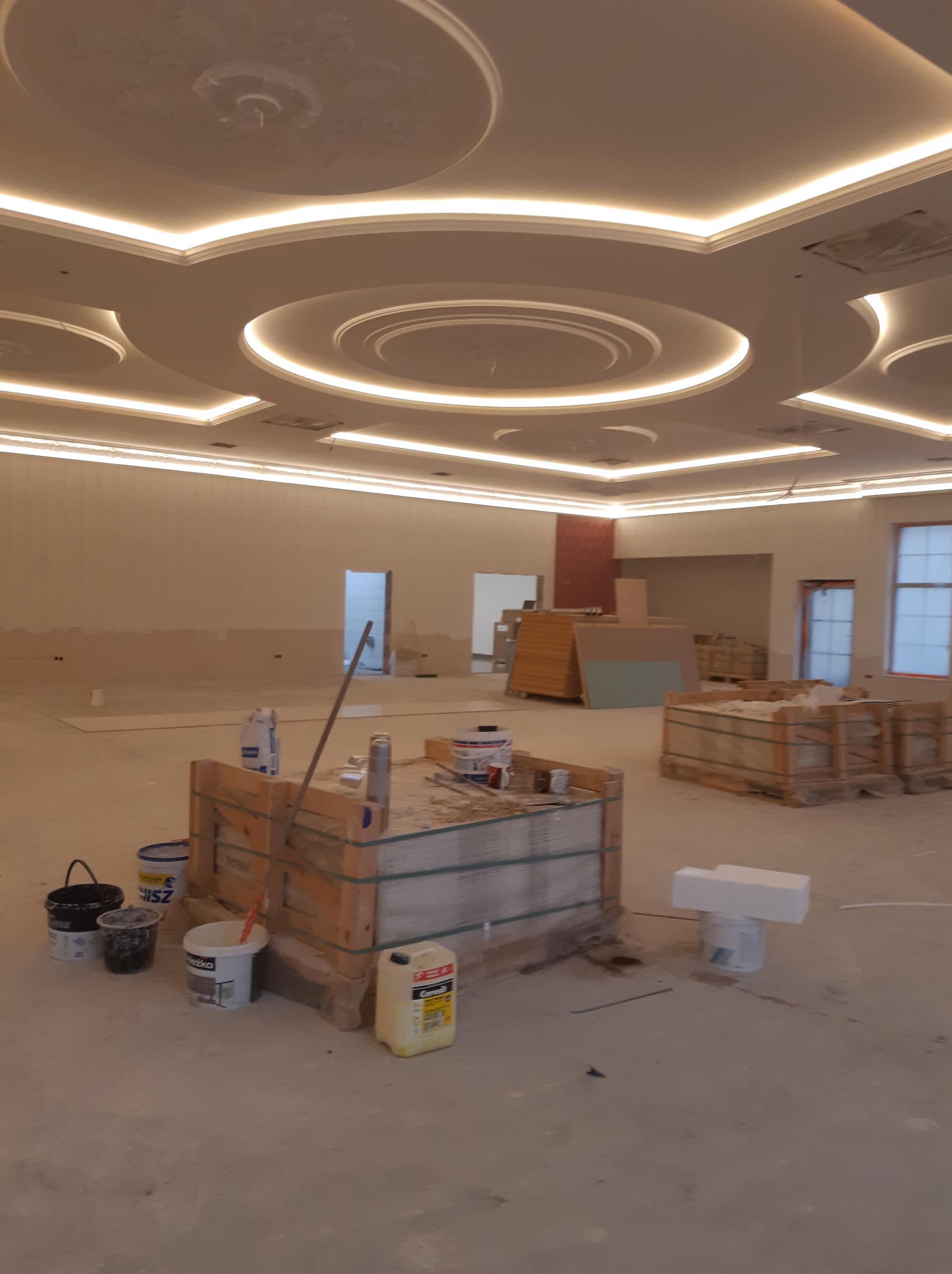 Wnętrze w trakcie prac wykończeniowych: widoczne gładkie ściany, dekoracyjny sufit z oświetleniem LED, materiały budowlane w pojemnikach i paletach, przygotowane do dalszych działań.