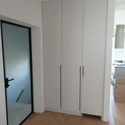 Biała szafa wnękowa z czarnymi, minimalistycznymi uchwytami, wkomponowana w jasne wnętrze z parkietem ułożonym we wzór jodełki, widoczna obok szklanych drzwi z czarną ramą.