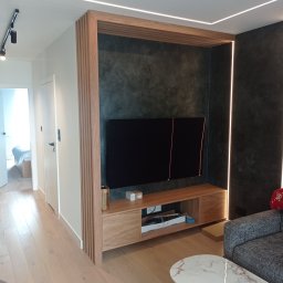 Karol Kieroński - Nowoczesny salon z drewnianą zabudową ściany TV, oświetleniem LED i blatem z drewna, widok na sypialnię w tle.