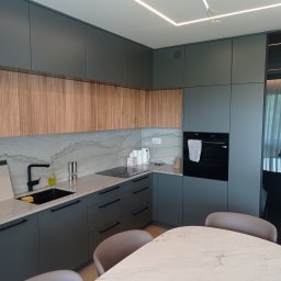 Karol Kieroński - Nowoczesna kuchnia z szarymi szafkami, drewnianymi frontami górnymi, marmurowym blatem i czarną armaturą, oświetlona ledowym paskiem pod sufitem. Widoczny fragment stołu z marmurowym blatem...