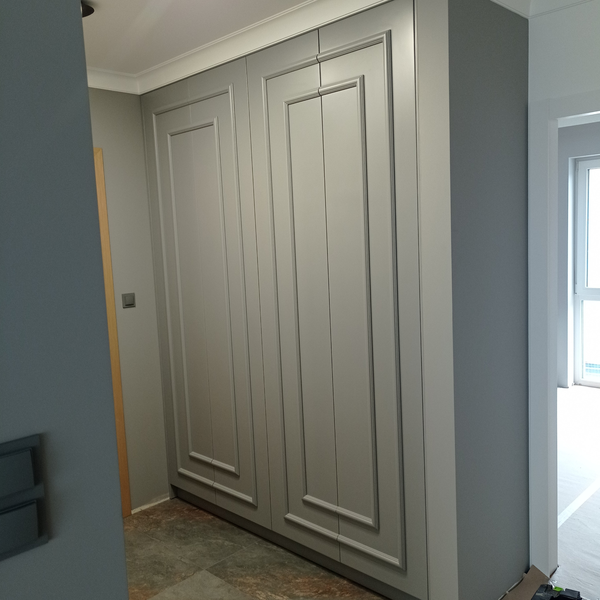 Szara szafa wnękowa z dekoracyjnymi frezowaniami na frontach, dopasowana kolorystycznie do ścian i listew przypodłogowych w nowoczesnym wnętrzu.