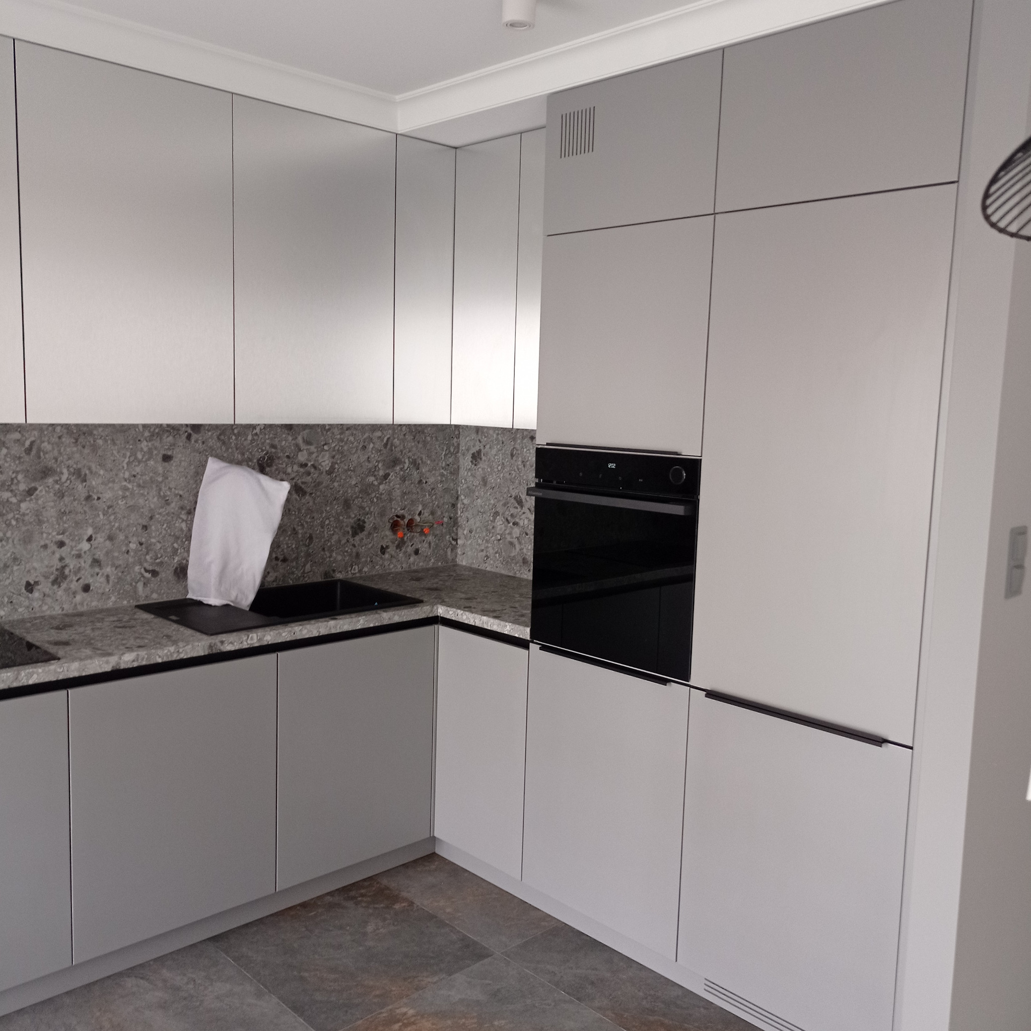 Nowoczesna kuchnia w odcieniach szarości z blatem imitującym kamień, z widocznym fragmentem instalacji wodnej i białym workiem w zlewie.