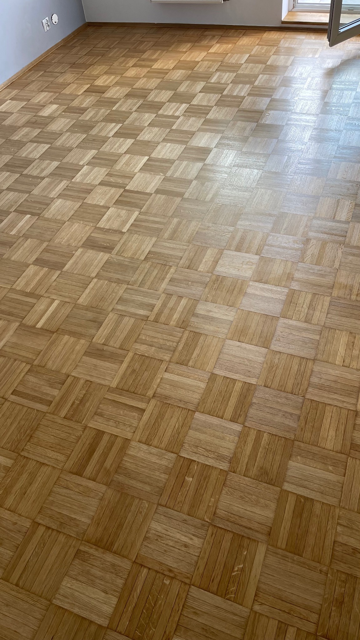 Parkiet ułożony we wzór szachownicy z naturalnego drewna dębowego, widoczny fragment ściany z gniazdkami elektrycznymi i okno w tle.