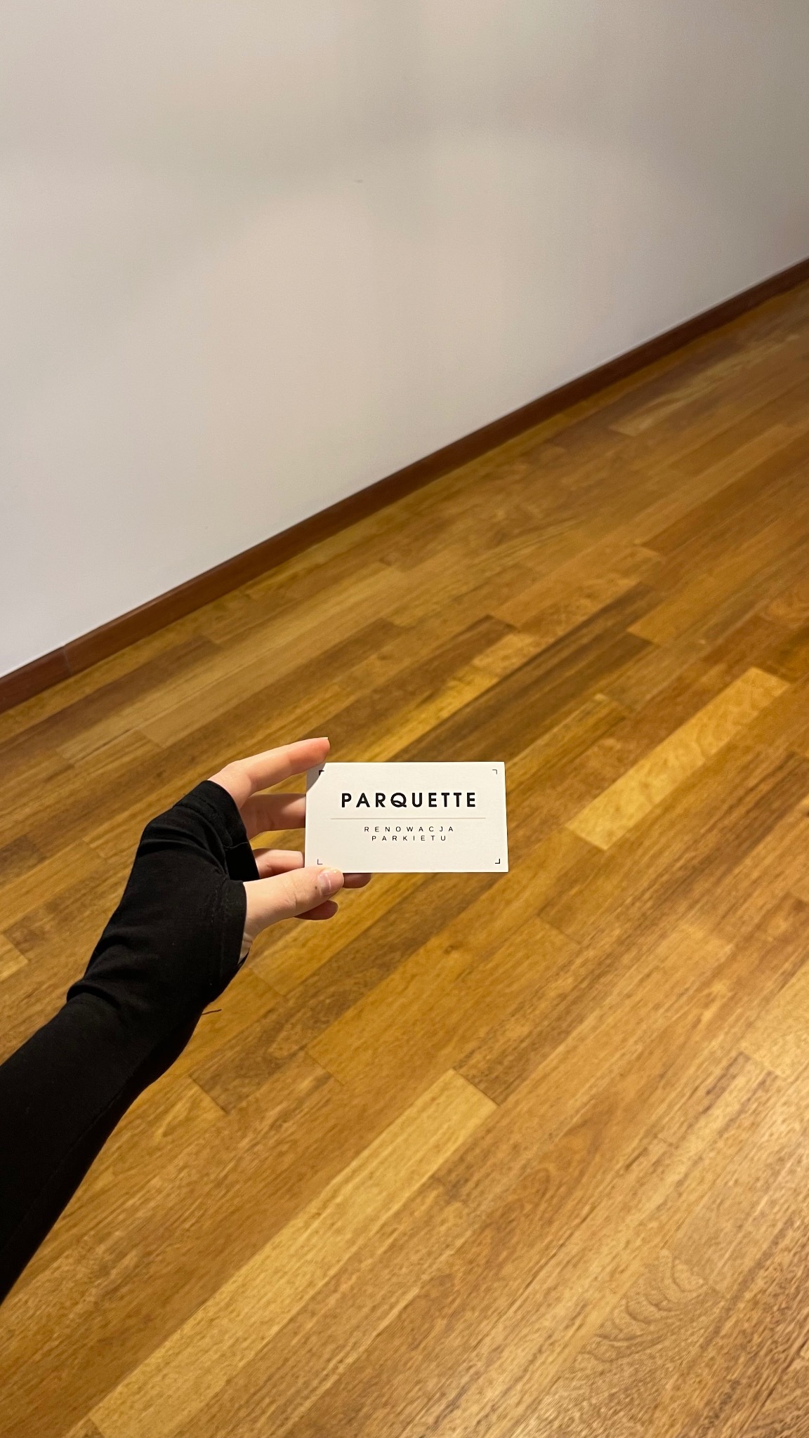 Dłoń w czarnej rękawiczce trzyma wizytówkę z napisem 'PARQUETTE Renowacja Parkietu' na tle parkietu z jasnego drewna i białej ściany.