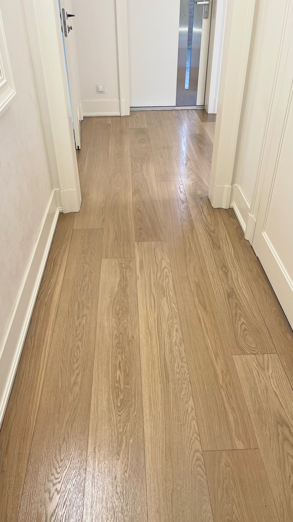 Jasny parkiet z wyraźnym usłojeniem, ułożony wzdłuż korytarza z białymi drzwiami i listwami przypodłogowymi.