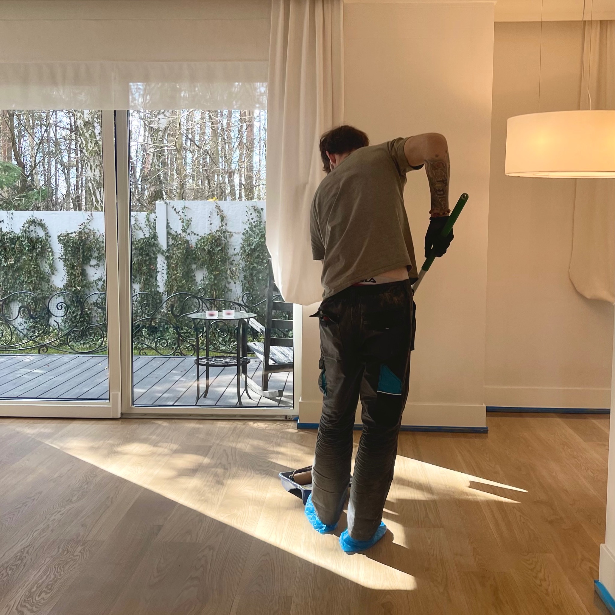 Parkieciarz w niebieskich ochraniaczach na buty, z wałkiem malarskim, wykańcza polerowanie nowej, jasnej drewnianej podłogi w pokoju z widokiem na taras i ogród.