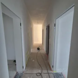 Korytarz po gładziowaniu i malowaniu na biało, widoczne zabezpieczenia podłogi i futryn. Na podłodze leżą kable od szlifierki.