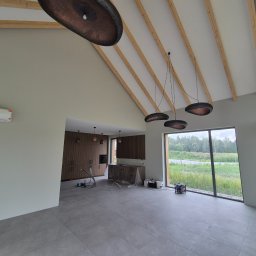 FAKTORIA GROUP SPÓŁKA Z OGRANICZONĄ ODPOWIEDZIALNOŚCIĄ - Wnętrze domu z drewnianą konstrukcją dachu i szarymi płytkami. Widoczne lampy wiszące, okno z widokiem na zieleń i sprzęt malarski. Ściany świeżo pomalowane na jasny odcień.