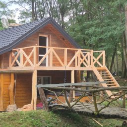 FAKTORIA GROUP SPÓŁKA Z OGRANICZONĄ ODPOWIEDZIALNOŚCIĄ - Drewniany dom z balkonem w trakcie budowy, widoczny mostek z gałęzi i pracownik przy balustradzie. Otoczenie leśne, pochmurne niebo.