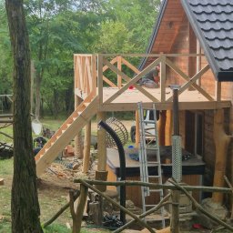 FAKTORIA GROUP SPÓŁKA Z OGRANICZONĄ ODPOWIEDZIALNOŚCIĄ - Drewniany domek z tarasem w trakcie budowy. Widoczne schody, balustrada z krzyżowych belek i drabina. W tle zieleń drzew i częściowo widoczna osoba pracująca na tarasie.