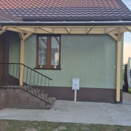 FAKTORIA GROUP SPÓŁKA Z OGRANICZONĄ ODPOWIEDZIALNOŚCIĄ - Zadaszenie wejścia do domu z jasnego drewna, widok z zewnątrz. Elementy konstrukcyjne złączone śrubami. W tle ściana domu z oknem i skrzynką pocztową.