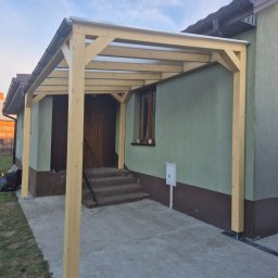 FAKTORIA GROUP SPÓŁKA Z OGRANICZONĄ ODPOWIEDZIALNOŚCIĄ - Drewniana pergola z przezroczystym zadaszeniem przy domu z zieloną elewacją. Widoczne schody wejściowe i skrzynka pocztowa. Ujęcie z dołu, niebieskie niebo.