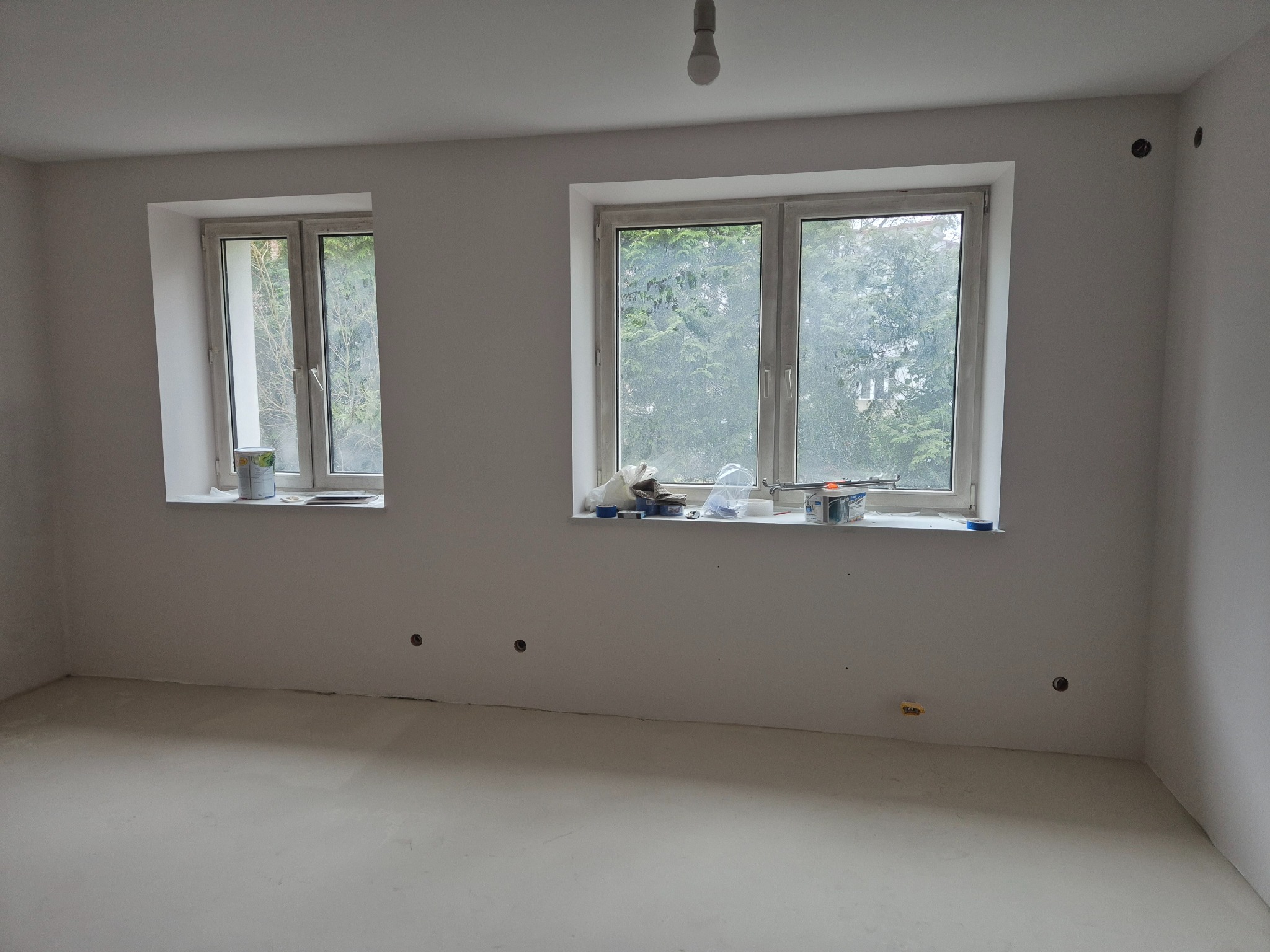 Pomalowane na biało wnętrze pokoju z dwoma oknami, na parapetach puszki z farbą i narzędzia malarskie, widoczne puste otwory na gniazdka elektryczne w ścianie.