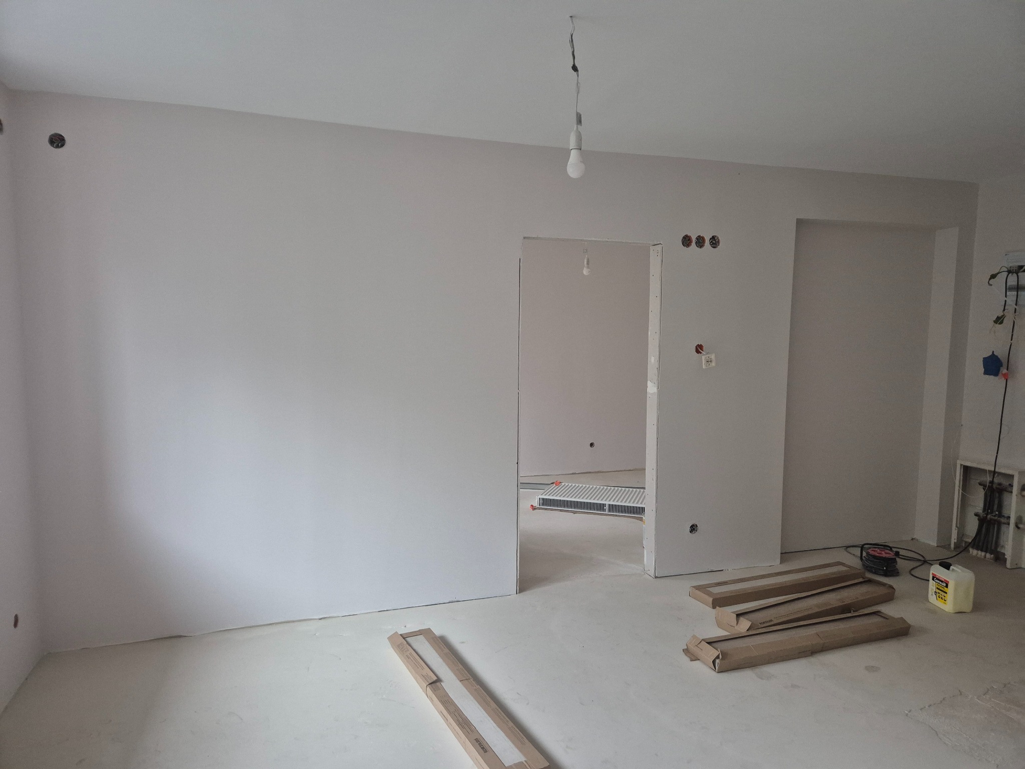 Wnętrze w trakcie prac wykończeniowych: świeżo pomalowane ściany na biało, widoczne puszki elektryczne, przygotowane do montażu listwy przypodłogowe w kartonowych opakowaniach, zwisająca żarówka...