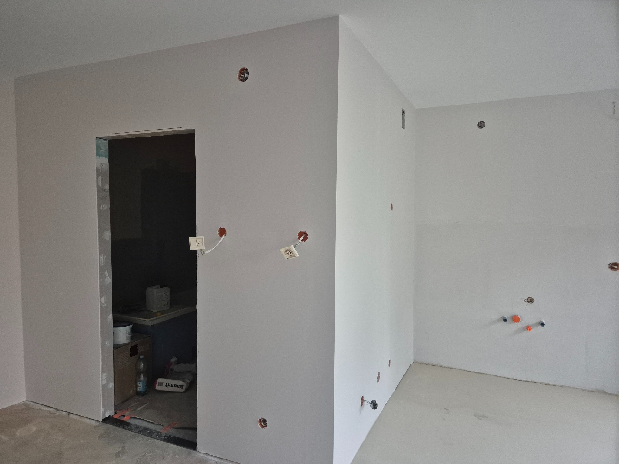 Wnętrze w trakcie prac wykończeniowych: świeżo pomalowane ściany w szarym odcieniu, widoczne puszki elektryczne i instalacja wodna, materiały budowlane w przejściu do sąsiedniego pomieszczenia.