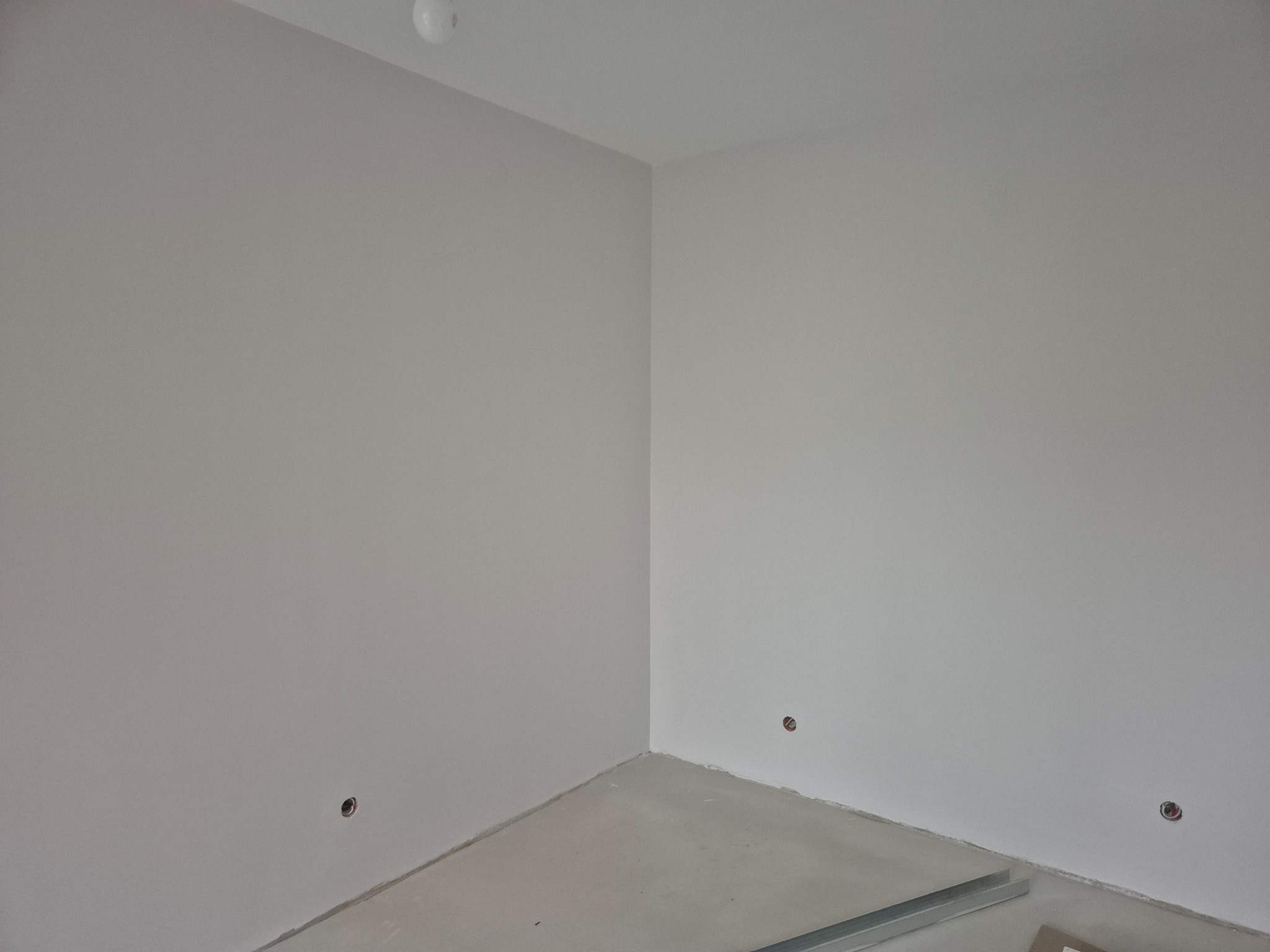 Wnętrze pokoju po szpachlowaniu ścian na gładko, widoczne otwory na gniazdka elektryczne i surowa podłoga z fragmentami profili.