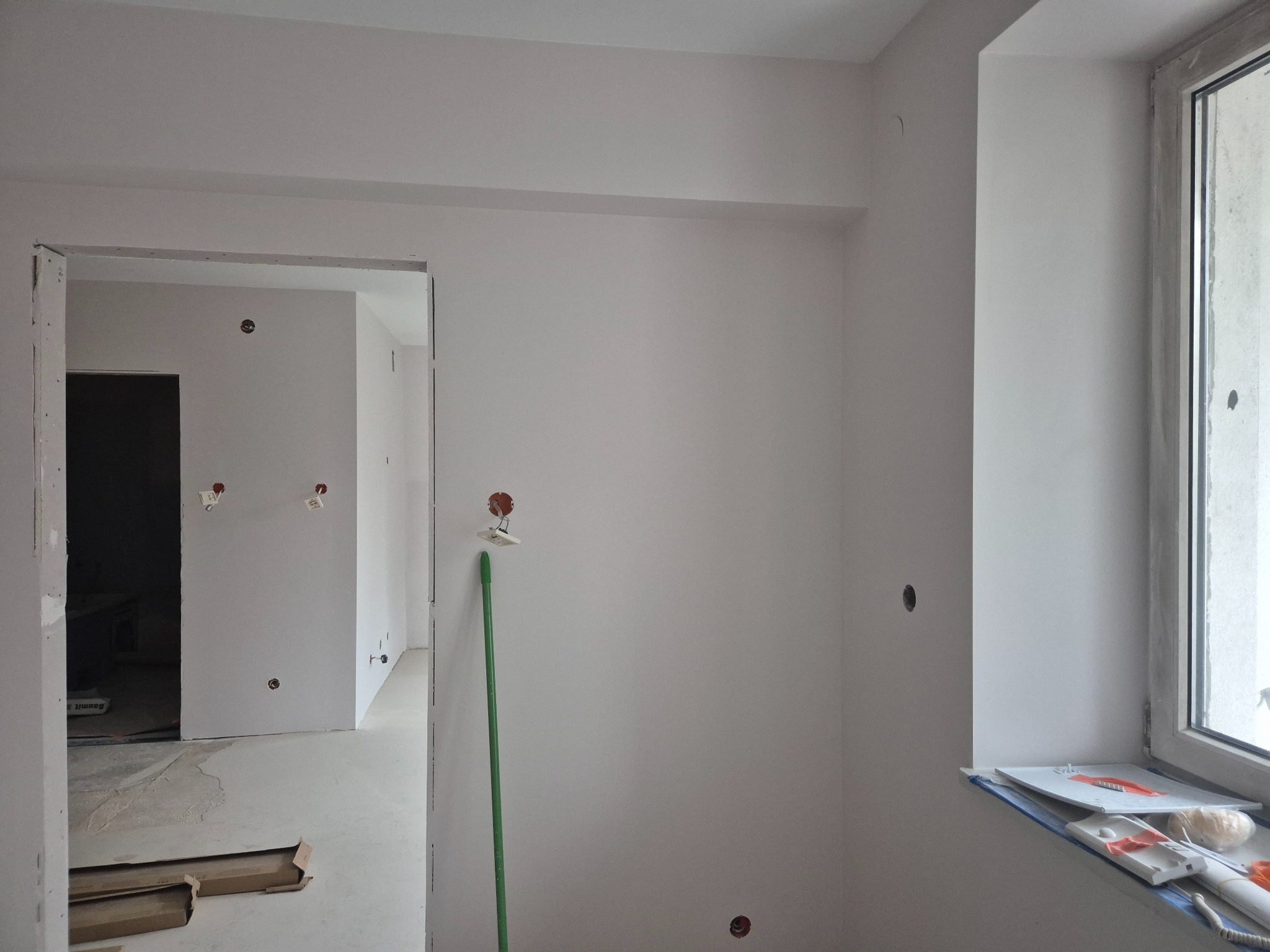 Wnętrze w trakcie prac wykończeniowych w Mszczonowie, z widocznymi otworami na gniazdka elektryczne w świeżo pomalowanej ścianie, zielona rurka instalacyjna przy ścianie, podłoga przygotowana...