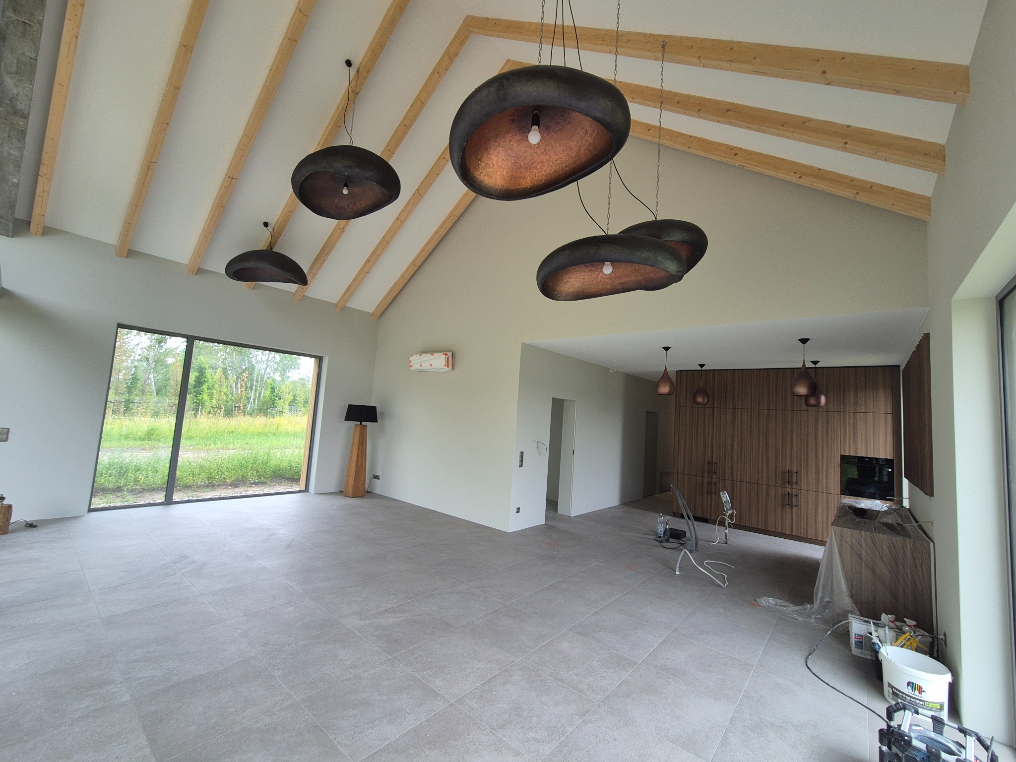 Jasne wnętrze domu z drewnianymi belkami stropowymi i nowoczesnymi lampami wiszącymi. Duże okno z widokiem na zieleń. Prace wykończeniowe w toku, widoczne narzędzia.