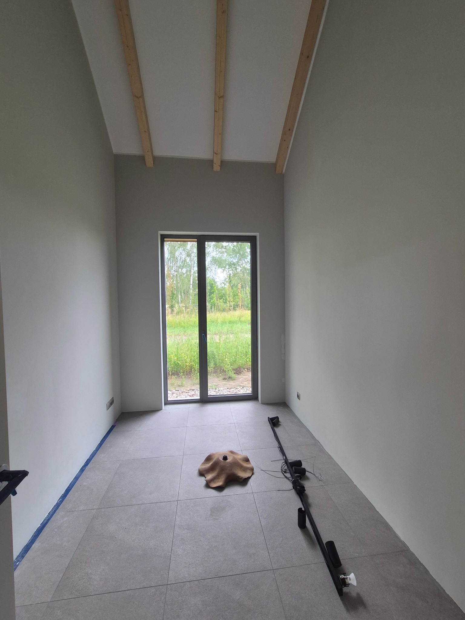 Jasne wnętrze po malowaniu ścian na szaro, z drewnianymi belkami na suficie, szarą podłogą i widokiem na zieleń za oknem. Taśma malarska przy podłodze.