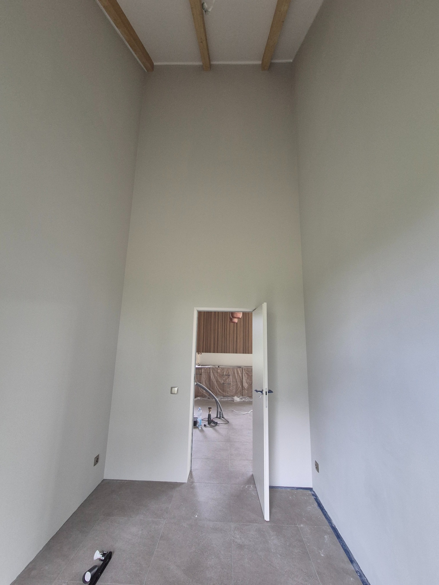 Wysokie, pomalowane na szaro wnętrze z drewnianymi belkami stropowymi, szarą podłogą i otwartymi drzwiami, przez które widać zakryte meble. Widoczne gniazdka elektryczne.