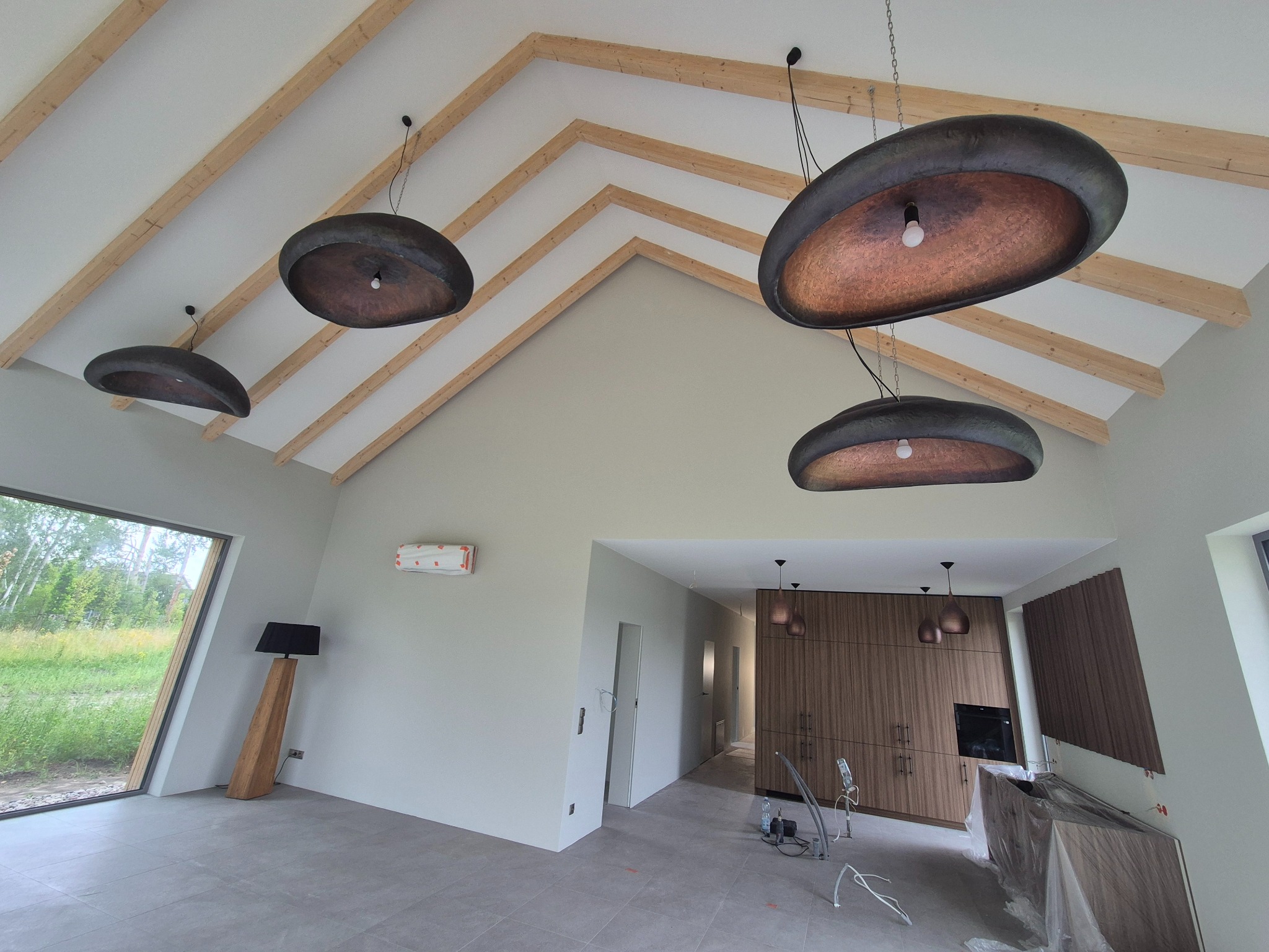 Nowoczesne wnętrze domu ze skośnym sufitem i drewnianymi belkami. Widoczne designerskie lampy wiszące, szara podłoga i duże okno. Świeżo pomalowane ściany.