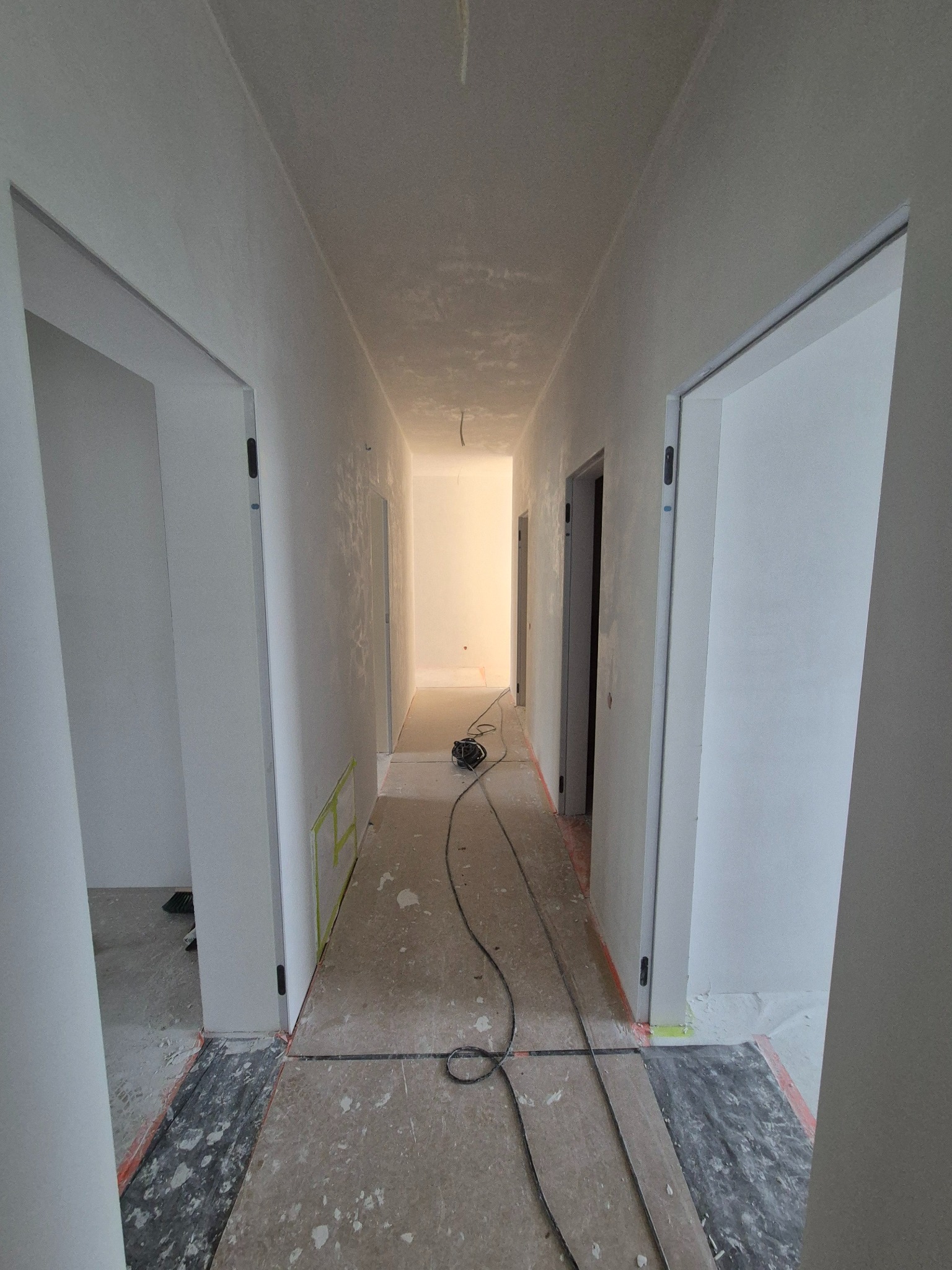 Korytarz po gładziowaniu i malowaniu na biało, widoczne zabezpieczenia podłogi i futryn. Na podłodze leżą kable od szlifierki.