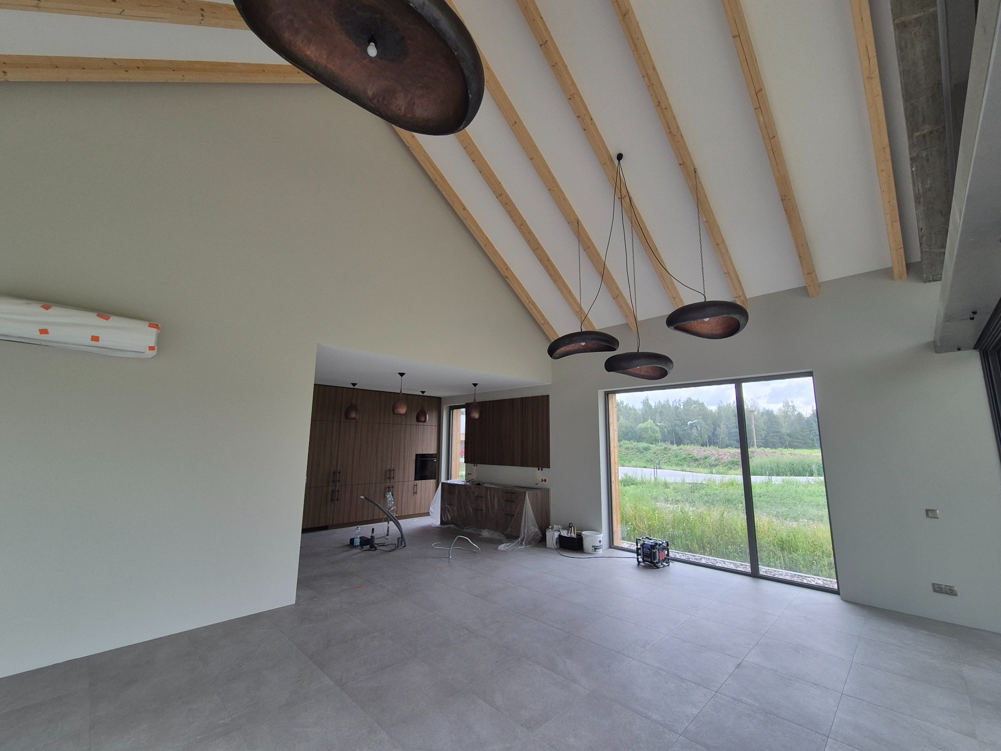 Wnętrze domu z drewnianą konstrukcją dachu i szarymi płytkami. Widoczne lampy wiszące, okno z widokiem na zieleń i sprzęt malarski. Ściany świeżo pomalowane na jasny odcień.