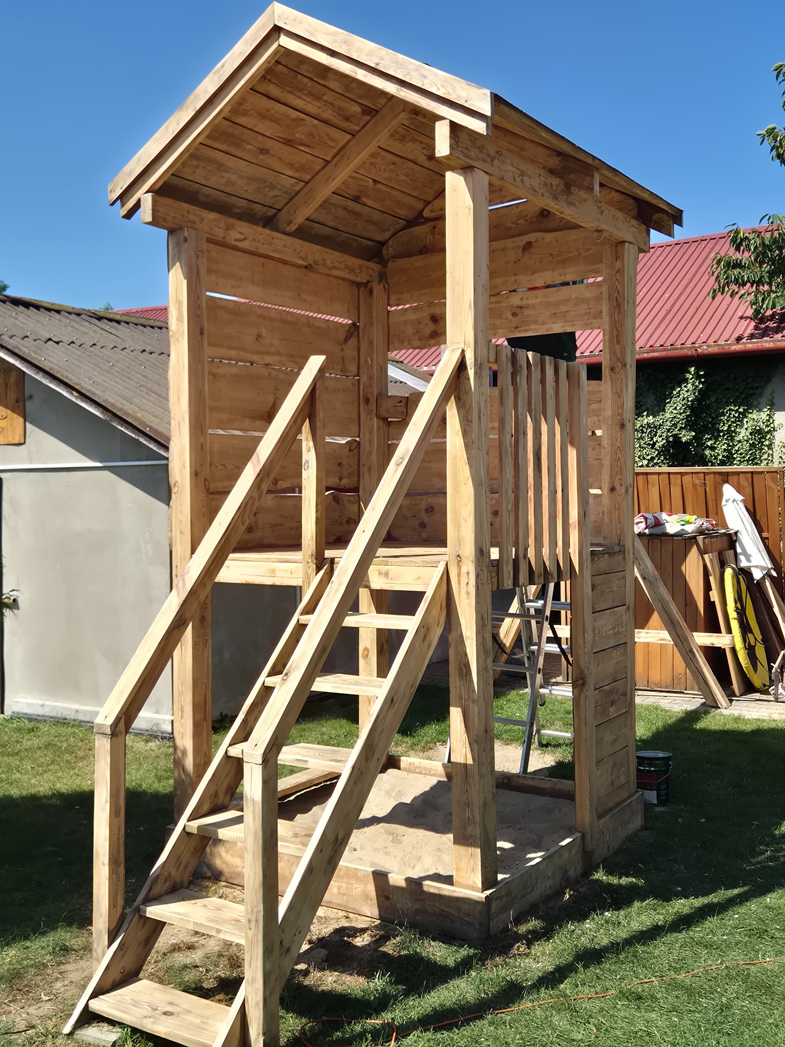 Drewniana konstrukcja placu zabaw z drabinką i zadaszeniem, w ogrodzie z trawą i zabudowaniami w tle, w słoneczny dzień. Widoczne detale konstrukcyjne i naturalne drewno.