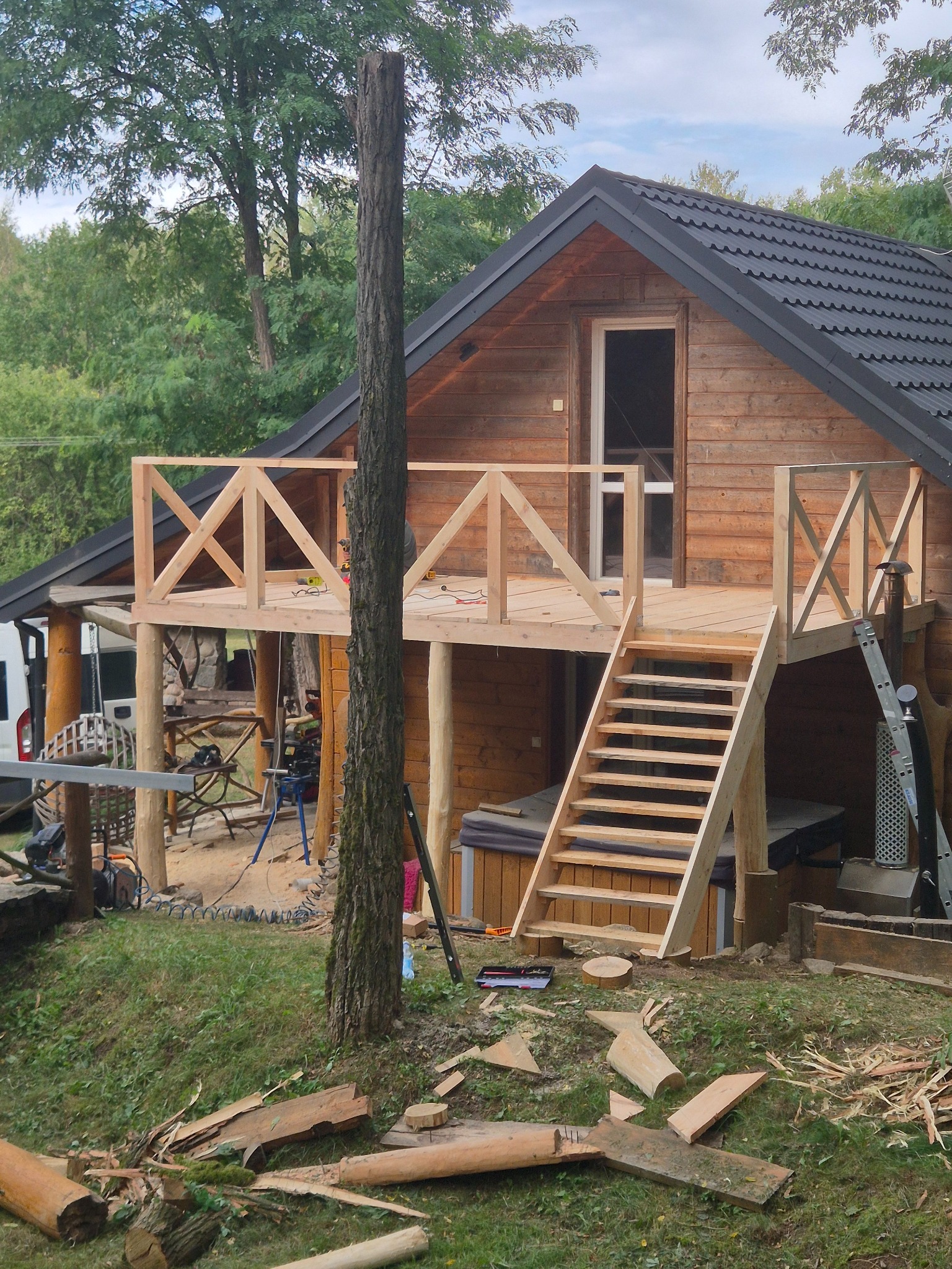 Drewniany domek z tarasem w trakcie budowy. Widoczne schody, balustrada z krzyżowymi elementami i narzędzia. Wokół drzewa i resztki drewna.