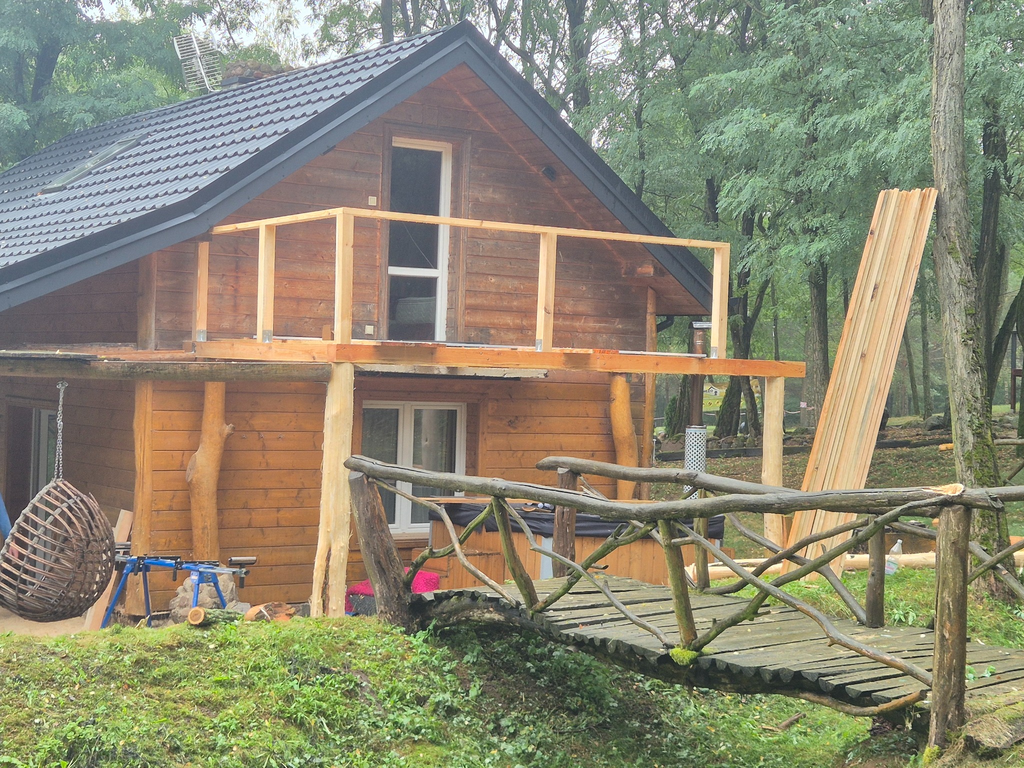 Drewniany domek z balkonem w trakcie budowy, widoczny mostek z bali, narzędzia i deski. Prace wykończeniowe w otoczeniu drzew.