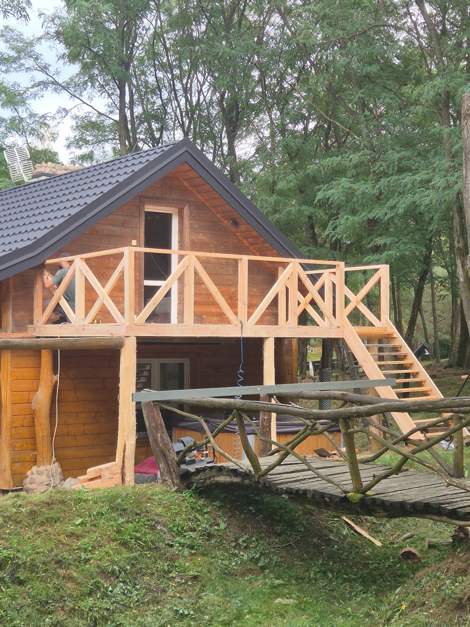 Drewniany dom z balkonem w trakcie budowy, widoczny mostek z gałęzi i pracownik przy balustradzie. Otoczenie leśne, pochmurne niebo.