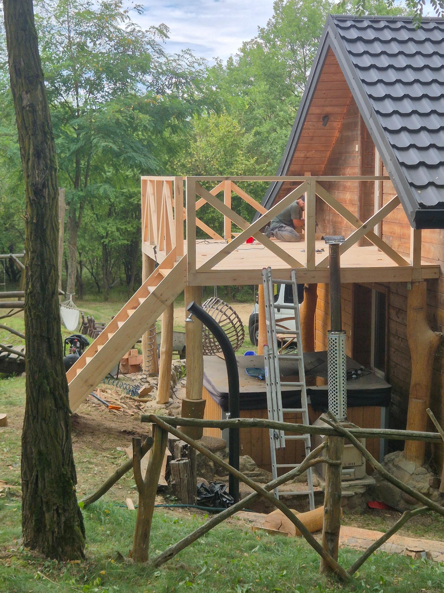 Drewniany domek z tarasem w trakcie budowy. Widoczne schody, balustrada z krzyżowych belek i drabina. W tle zieleń drzew i częściowo widoczna osoba pracująca na tarasie.