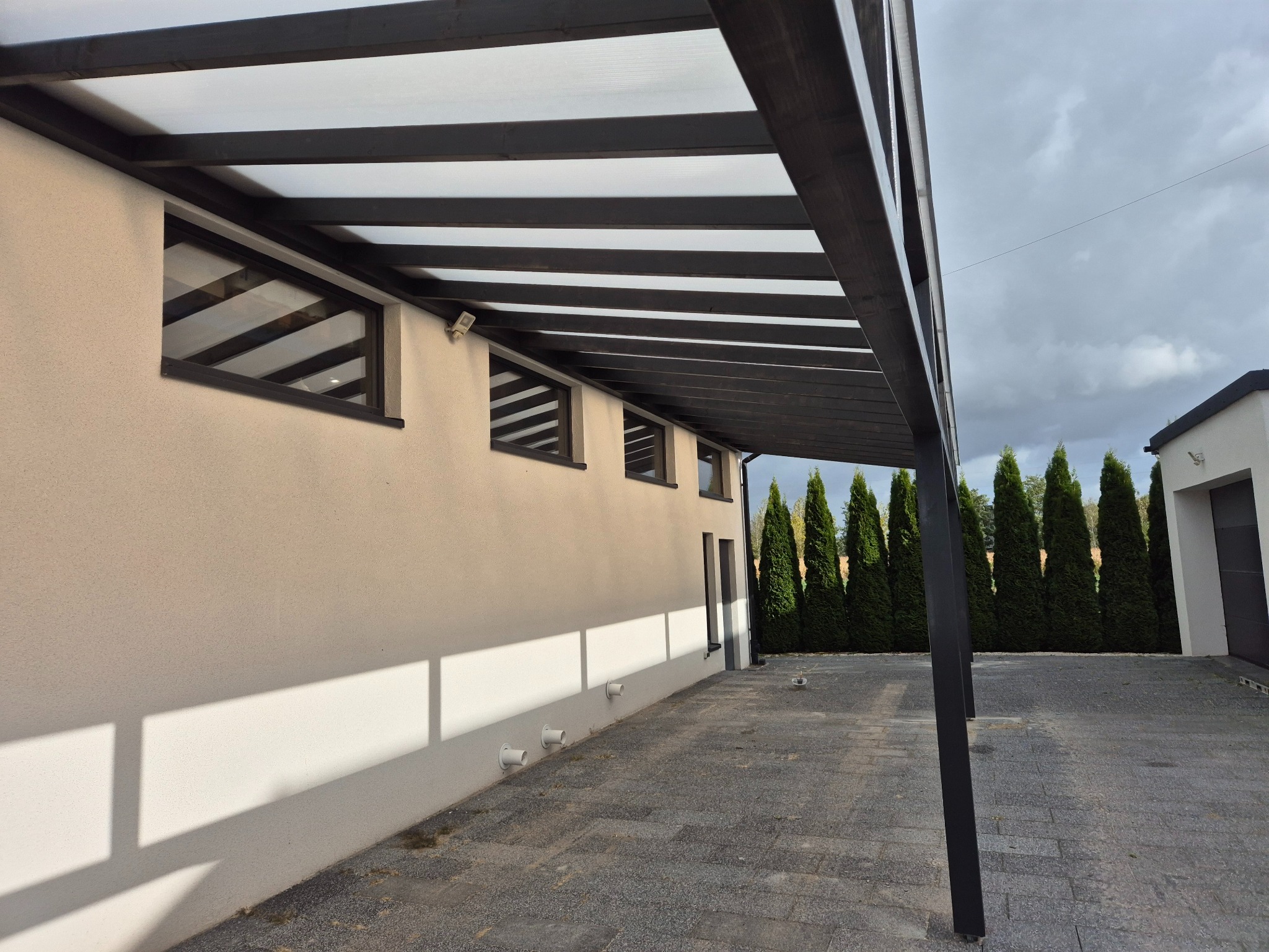 Nowoczesna pergola z ciemnego drewna przylegająca do jasnej ściany budynku z małymi oknami. Widoczne tuje w tle i kostka brukowa na podłożu.