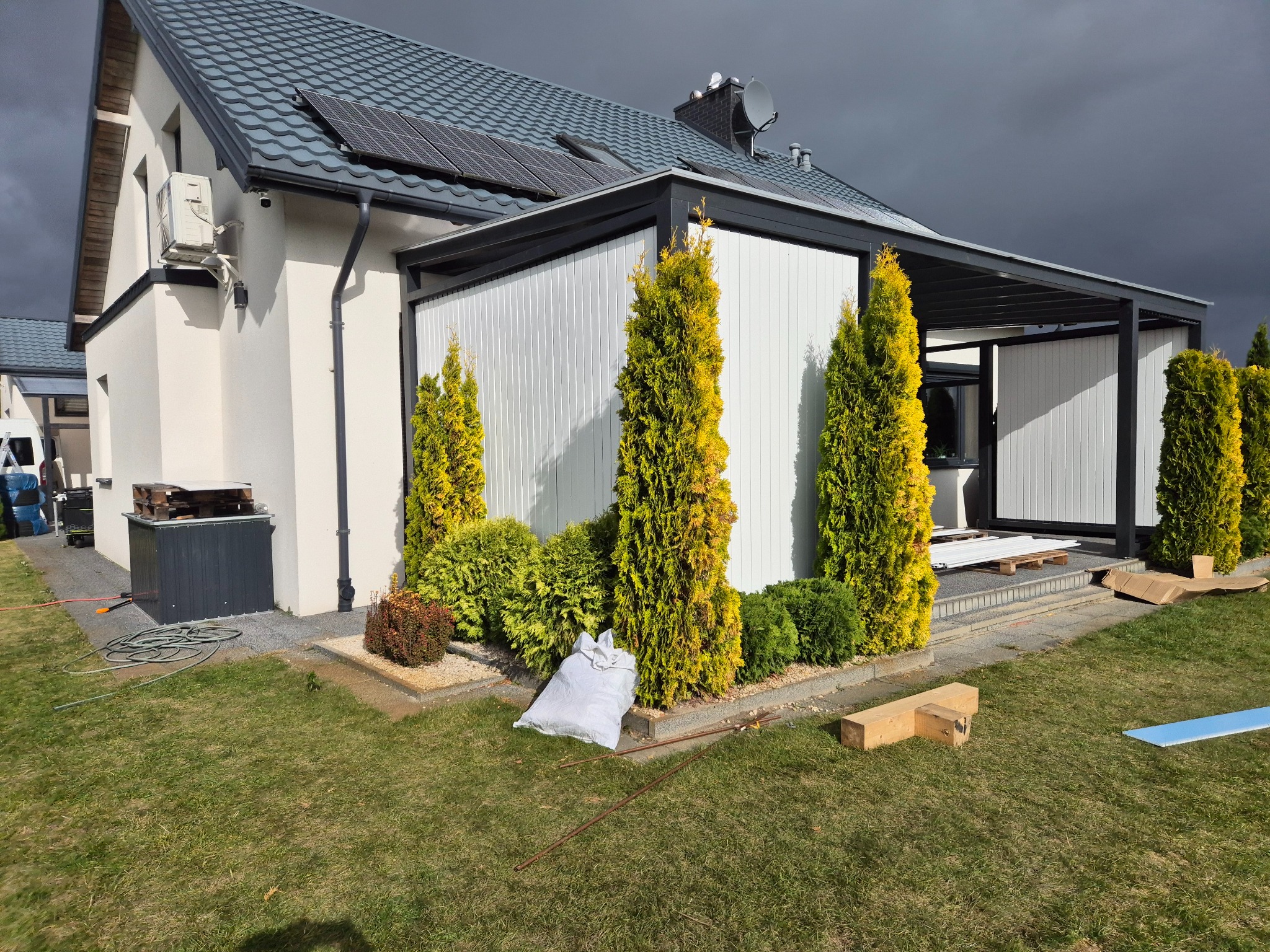 Nowoczesna pergola z białymi panelami i czarną ramą, otoczona zielenią. Widoczne elementy montażowe i narzędzia sugerują trwający montaż.