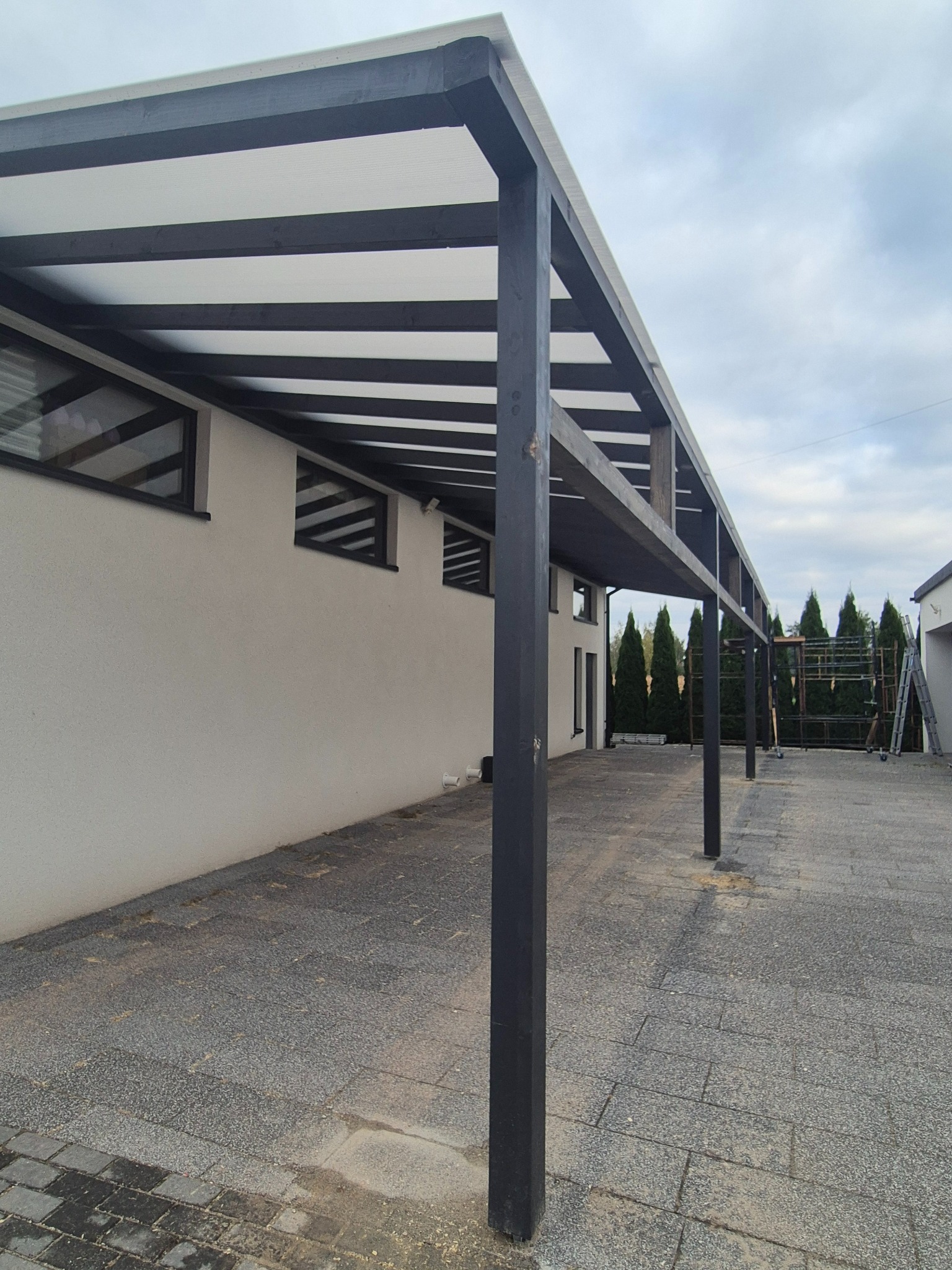 Nowoczesna, zadaszona wiata garażowa z czarną konstrukcją i białym dachem, przylegająca do budynku z małymi oknami. Widok z zewnątrz, kostka brukowa na podłożu.