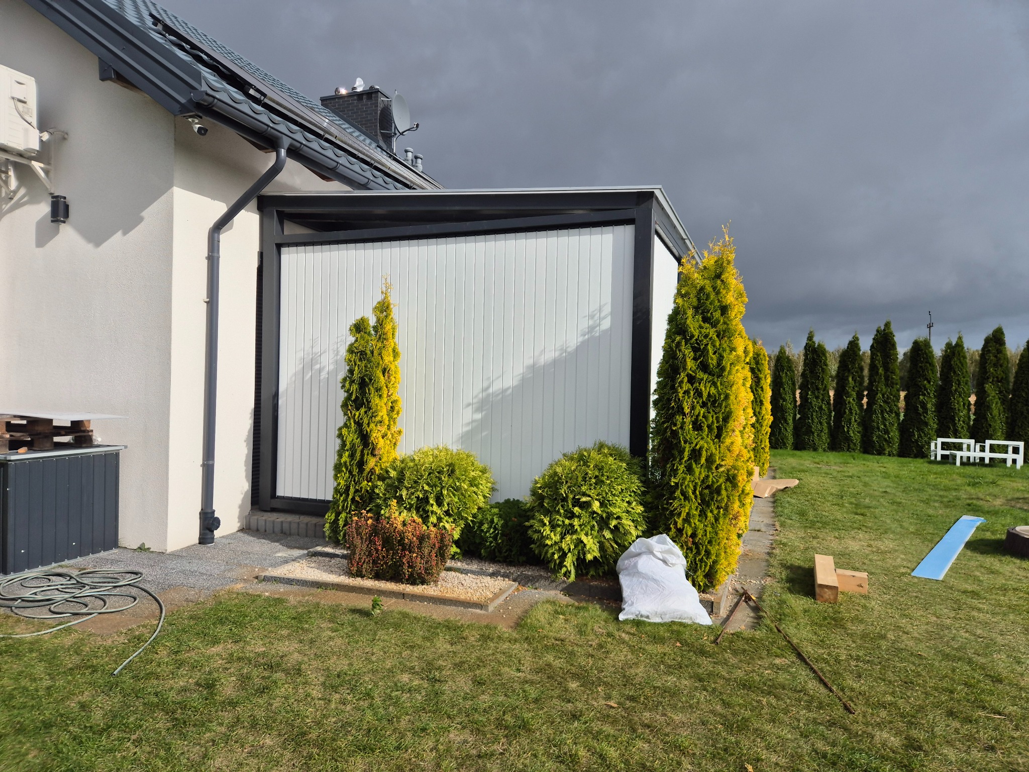 Biała elewacja domu z szarą pergolą i rzędem tuj na tle pochmurnego nieba. Widoczny fragment ogrodu z elementami wyposażenia.