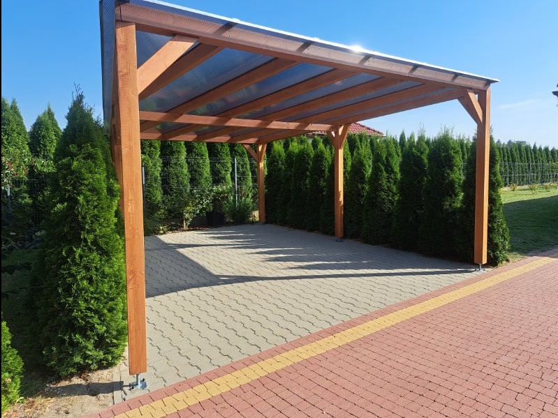 Drewniana wiata garażowa z przezroczystym zadaszeniem, wsparta na metalowych stopach, na tle żywopłotu z tui i kostki brukowej. Słoneczny dzień.