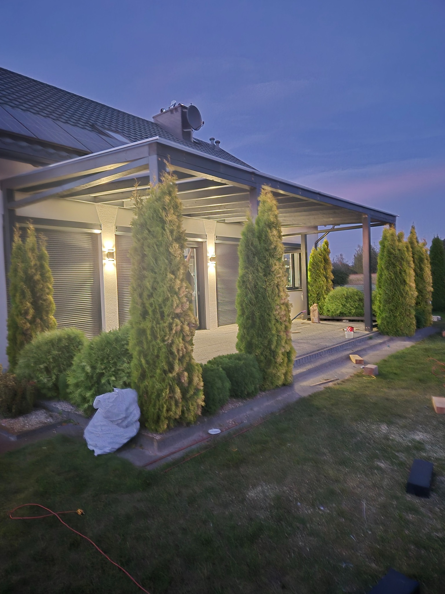 Nowoczesna pergola z oświetleniem i roletami zewnętrznymi, otoczona tujami i krzewami na tle domu z panelami słonecznymi, sfotografowana o zmierzchu.