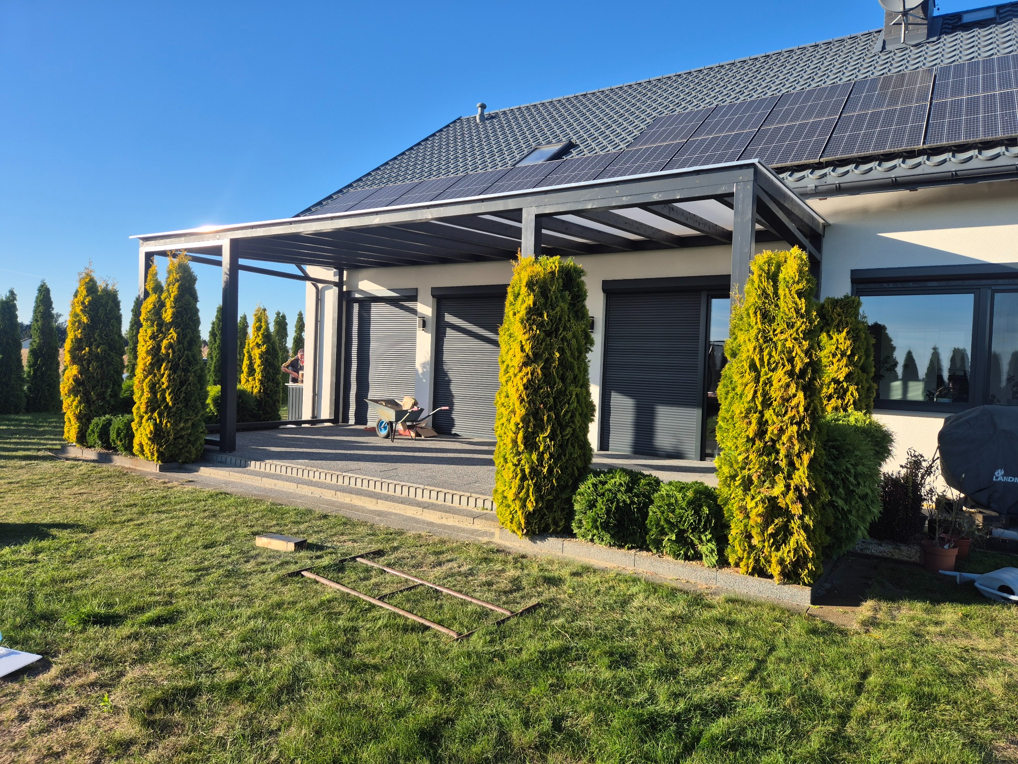 Nowoczesna pergola z zadaszeniem przy domu jednorodzinnym. Widoczne panele słoneczne na dachu, tuje i trawnik. Ujęcie w słoneczny dzień.