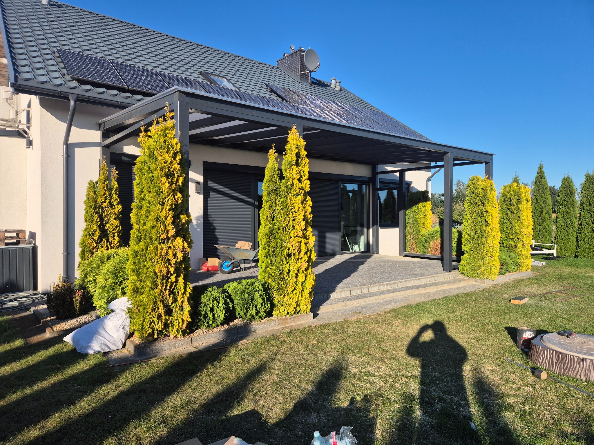 Nowoczesna pergola z szarymi roletami przy domu z panelami słonecznymi, otoczona tujami i zielonym trawnikiem w słoneczny dzień. Widoczny cień osoby.