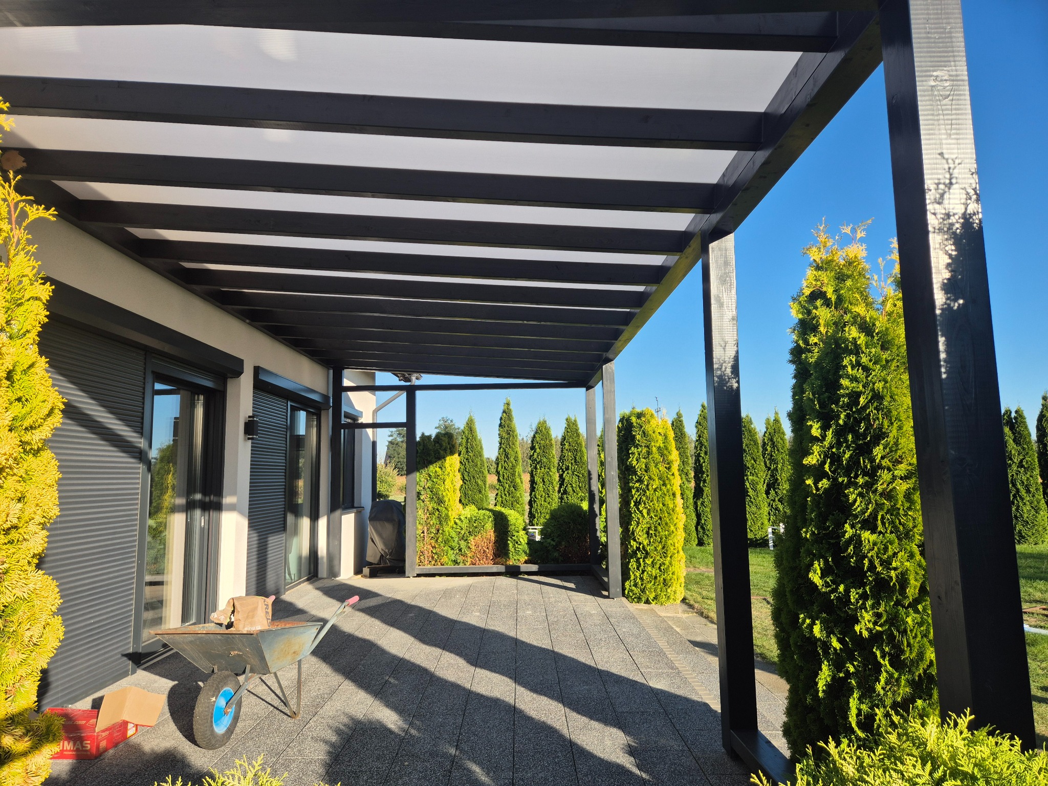 Nowoczesna pergola z czarnymi belkami i jasnym zadaszeniem, rzucająca cienie na szarą kostkę brukową. W tle rząd tuj i fragment budynku z szarymi roletami.